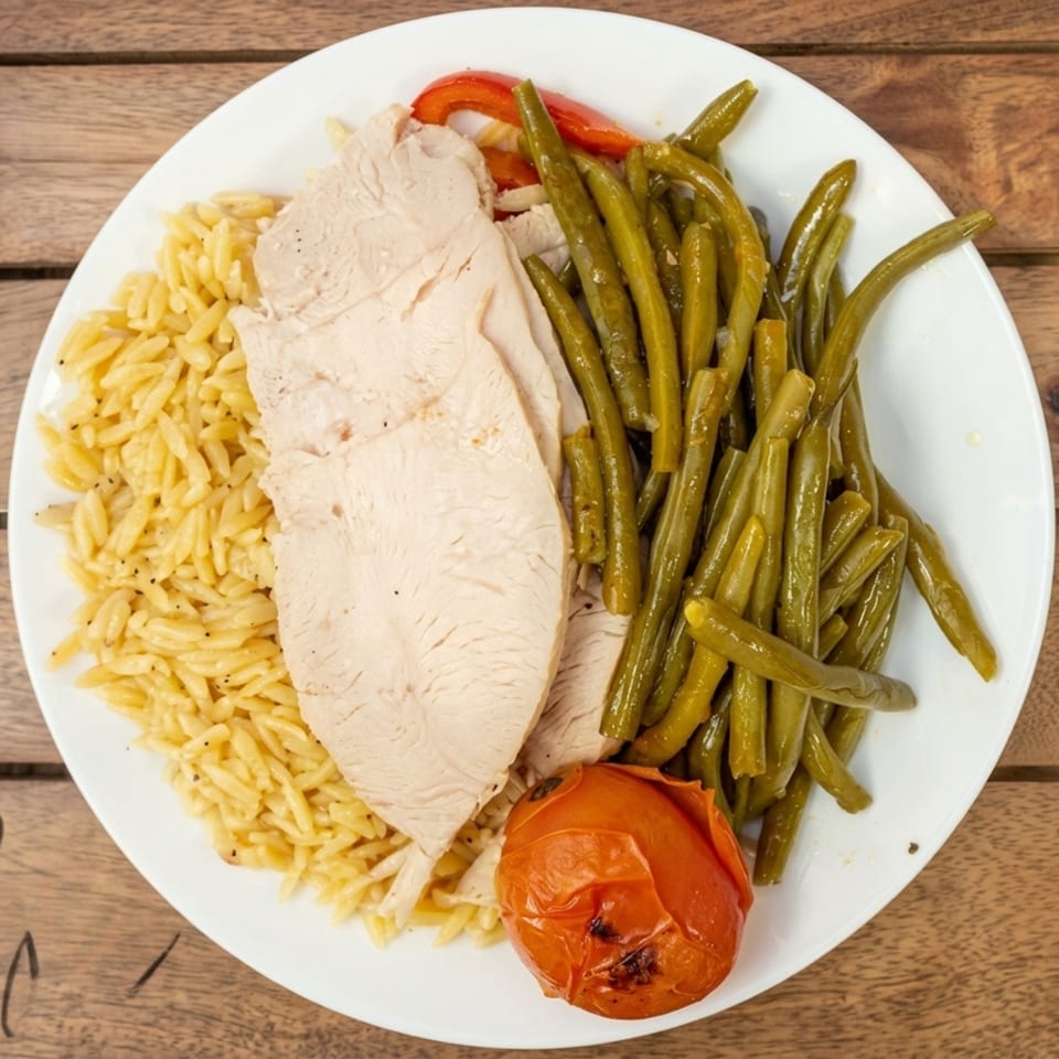 Platter with sliced turkey, orzo pasta, green beans, and a roasted tomato on a wooden table.