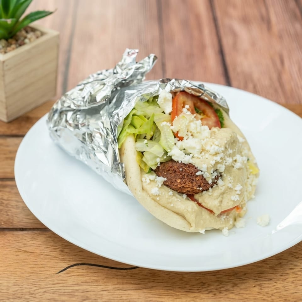 Falafel gyro with lettuce, tomatoes, feta, and hummus, partially wrapped in foil on a white plate.